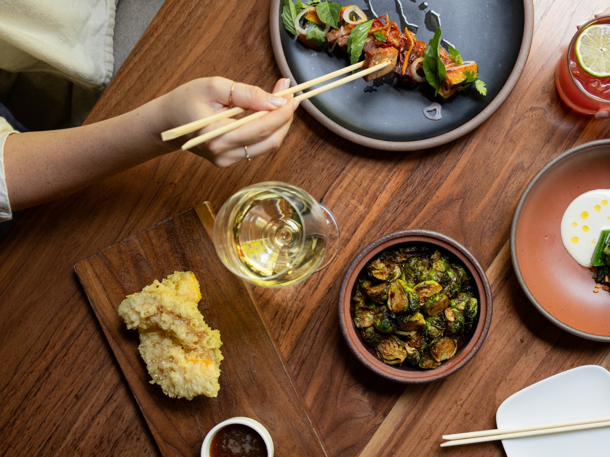 A close-up of a hand holding a pair of chopsticks surrounded by a table of food and drink.